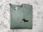 Folded military green fleece sweatshirt featuring a cute embroidered black and tan wiener dog on left chest on soft grey fur