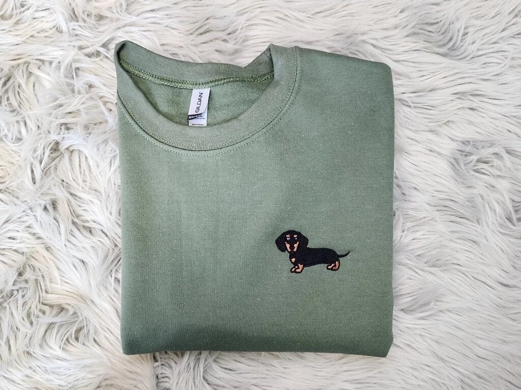 Dachshund Embroidered Crewneck Sweatshirt – Military Green Folded Display Folded military green fleece sweatshirt featuring a cute embroidered black and tan wiener dog on left chest on soft grey fur