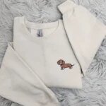Flat lay of sand beige crewneck sweater with delicate brown and white dachshund embroidery on left chest and open sleeve cuffs on textured fur