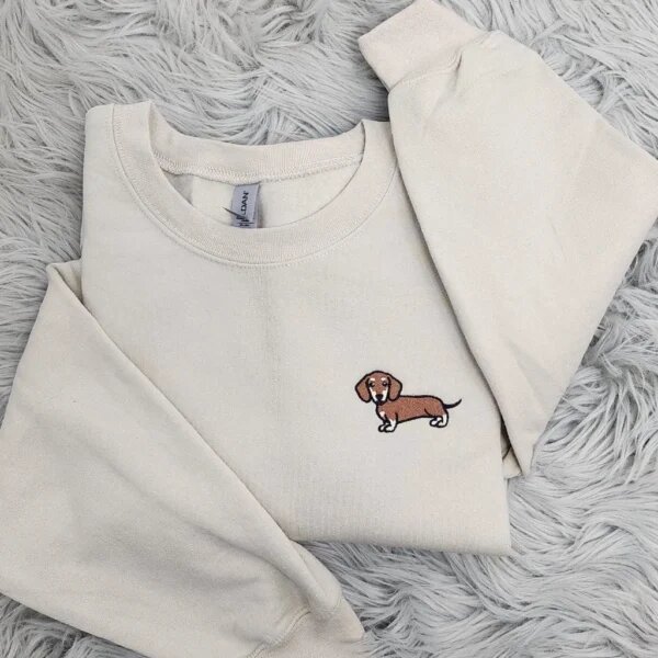 Flat lay of sand beige crewneck sweater with delicate brown and white dachshund embroidery on left chest and open sleeve cuffs on textured fur