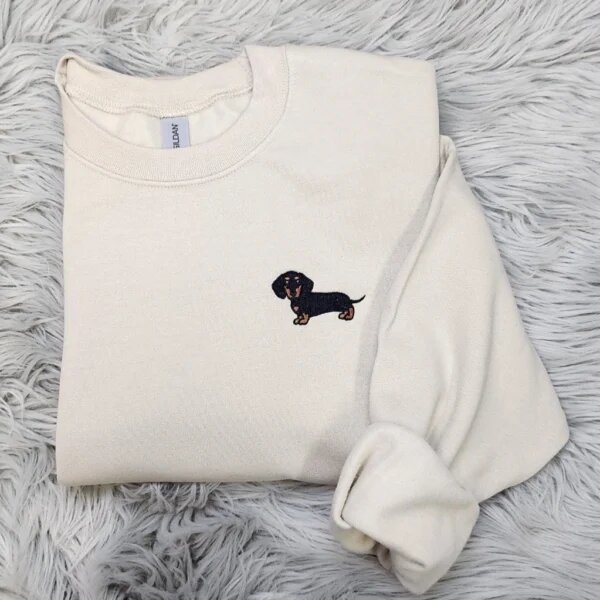 Folded sand beige fleece crewneck sweatshirt featuring detailed embroidered black and tan dachshund dog on the left chest resting on grey fur rug