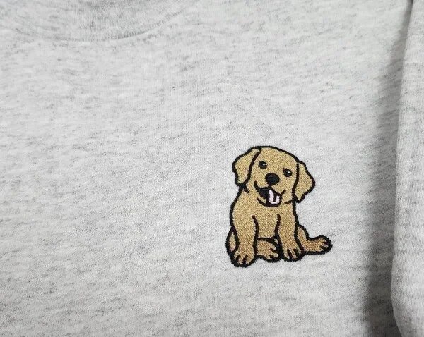 Macro close-up view of textured thread embroidery showing a golden labrador retriever puppy with pink tongue and black outline on heather grey sweatshirt fleece