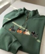 Forest green quarter zip sweater showcasing embroidered autumn dachshund lineup in spooky Halloween costumes on a sunny white table