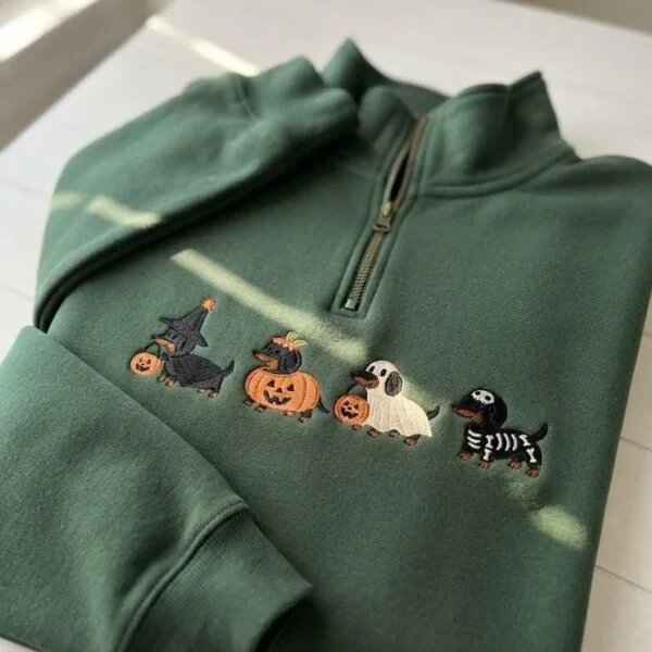 Forest green quarter zip sweater showcasing embroidered autumn dachshund lineup in spooky Halloween costumes on a sunny white table