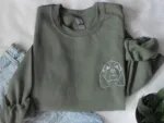 Folded olive green crewneck sweatshirt paired with light wash jeans featuring white thread embroidered floppy-eared spaniel dog face