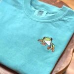 Folded seafoam teal embroidered frog t-shirt on a natural wooden table showcasing a detailed colorful mini tree frog with orange feet and vibrant green skin