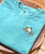 Folded seafoam teal embroidered frog t-shirt on a natural wooden table showcasing a detailed colorful mini tree frog with orange feet and vibrant green skin