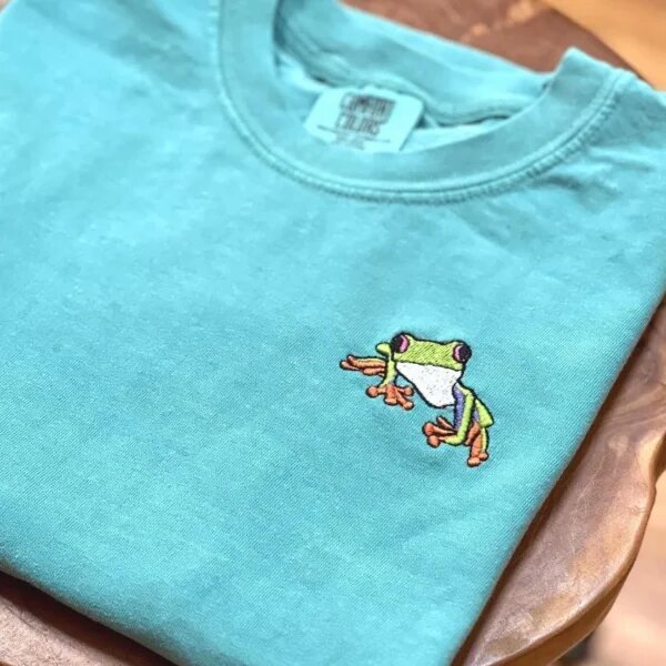 Folded seafoam teal embroidered frog t-shirt on a natural wooden table showcasing a detailed colorful mini tree frog with orange feet and vibrant green skin