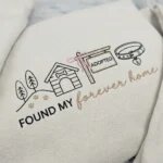 Macro close-up view of textured thread embroidery showing dog house, pink ribbon bow on adopted sign, collar, and cursive forever home lettering on sand fleece