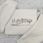 Folded sand beige fleece crewneck sweatshirt featuring embroidered dog house, pink bow adopted sign, collar, and Found My Forever Home script on grey plush fur rug