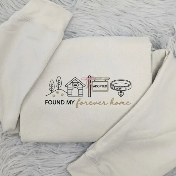 Folded sand beige fleece crewneck sweatshirt featuring embroidered dog house, pink bow adopted sign, collar, and Found My Forever Home script on grey plush fur rug