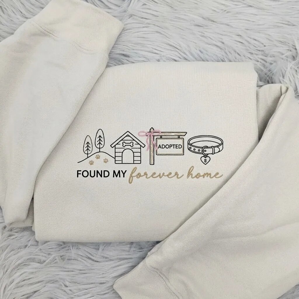 Found My Forever Home Embroidered Sweatshirt – Sand Beige Folded Flatlay Display Folded sand beige fleece crewneck sweatshirt featuring embroidered dog house, pink bow adopted sign, collar, and Found My Forever Home script on grey plush fur rug