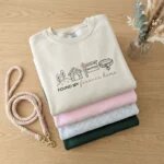 Folded sand beige embroidered sweatshirt with Found My Forever Home dog adoption design on top of pastel pink, heather grey, and forest green sweaters with braided dog leash