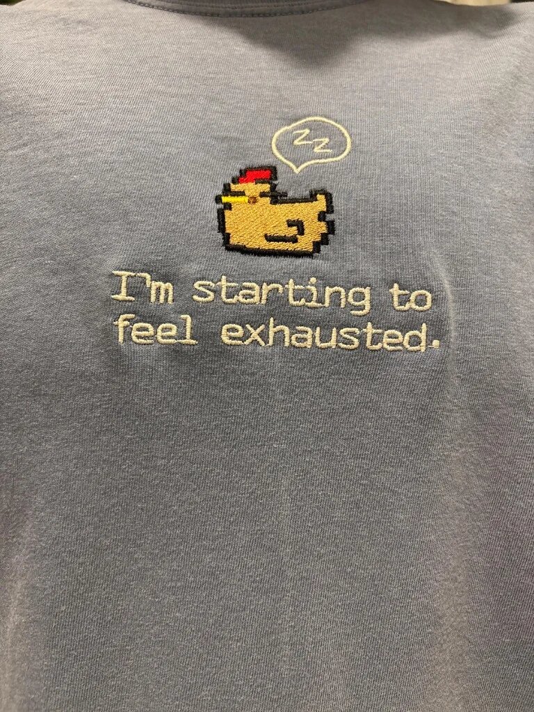 I'm Starting to Feel Exhausted Embroidered Apparel – Macro Thread Stitching Detail Detailed macro view of sleepy pixel chicken embroidery and crisp lettering on soft steel blue textured fabric