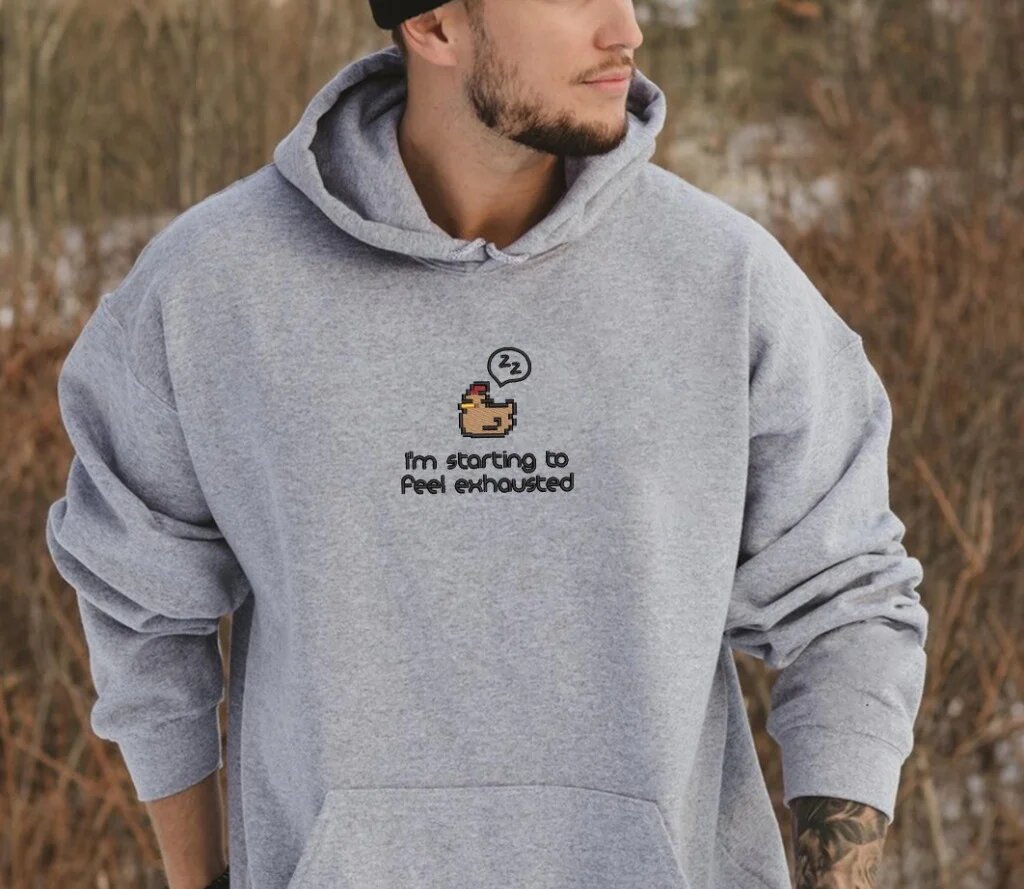 I'm Starting to Feel Exhausted Embroidered Hoodie – Heather Grey Lifestyle Model Model wearing heather grey embroidered pullover hoodie with sleepy chicken gamer motif outdoors in winter forest scenery