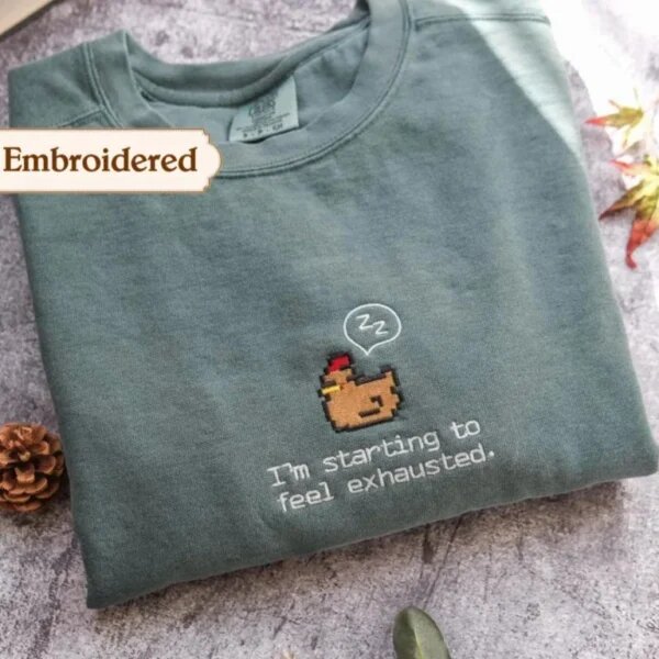Sage green embroidered crewneck sweatshirt with brown pixel art sleepy chicken and I'm starting to feel exhausted text on rustic slate with pinecone and autumn leaves