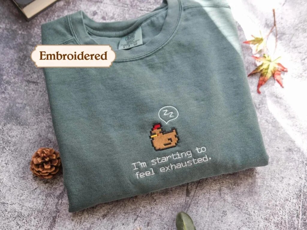 I'm Starting to Feel Exhausted Embroidered Sweatshirt – Sage Green Crewneck Flatlay Sage green embroidered crewneck sweatshirt with brown pixel art sleepy chicken and I'm starting to feel exhausted text on rustic slate with pinecone and autumn leaves