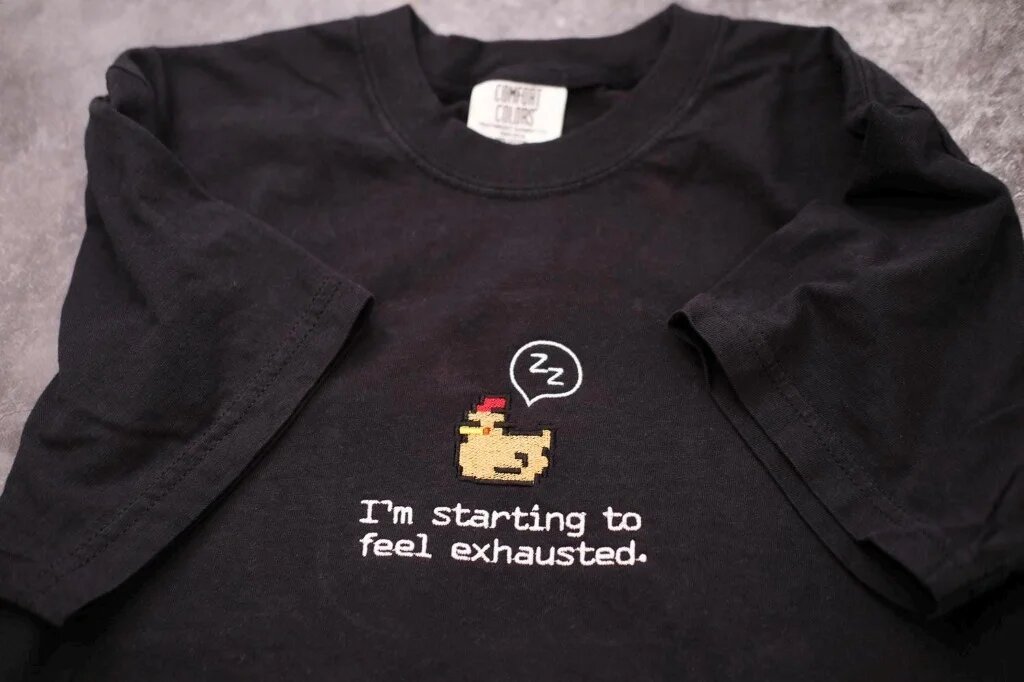 I'm Starting to Feel Exhausted Embroidered T-Shirt – Classic Black Flatlay Classic black embroidered cotton t-shirt with detailed brown pixel chicken and I'm starting to feel exhausted quote across chest