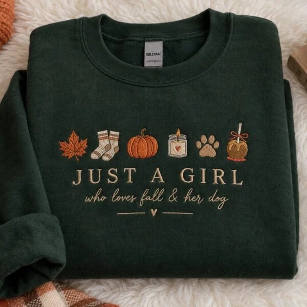 Cozy flat lay display of forest green embroidered fall sweatshirt resting on white sherpa fleece with plaid blanket, scented candle, and pumpkin decor