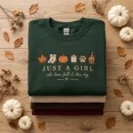 Folded forest green fleece sweatshirt embroidered with Just a Girl Who Loves Fall and Her Dog and autumn icons on stack of seasonal sweaters with white pumpkins on wood