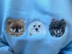Close-up macro detail of realistic textured embroidery showing three fluffy small dog portraits stitched on light blue fleece fabric