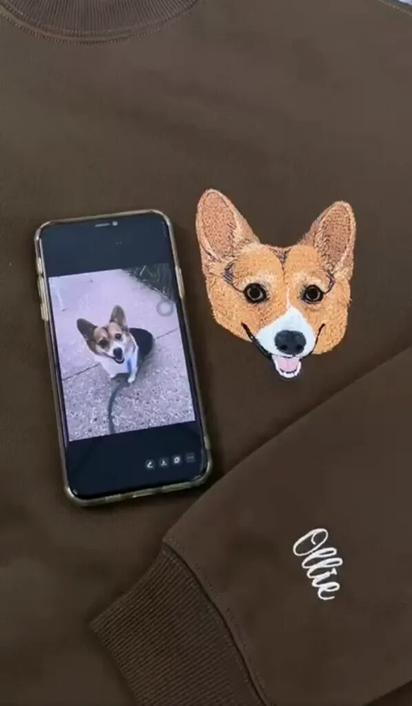 Phone showing a corgi photo beside the finished corgi portrait on a brown sweatshirt with the name Ollie stitched on the sleeve