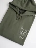 Folded olive green hoodie with drawstring featuring custom white thread embroidered cat face with whiskers and personalized pet name Abed