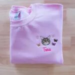 Folded light pink crewneck sweatshirt featuring custom embroidered tabby cat face named Tuna with floral daisies and butterflies on chest