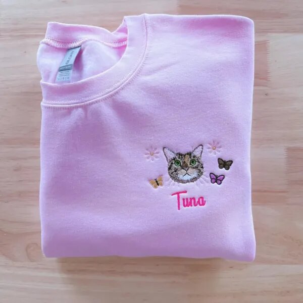 Folded light pink crewneck sweatshirt featuring custom embroidered tabby cat face named Tuna with floral daisies and butterflies on chest