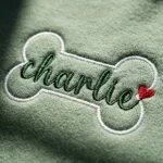 Extreme close-up macro view of dense high-sheen satin stitch cursive name embroidered across white thread dog bone outline on sand fleece