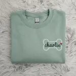 Folded oatmeal sand beige fleece crewneck sweatshirt featuring custom embroidered white dog bone outline with navy cursive pet name on grey fur rug