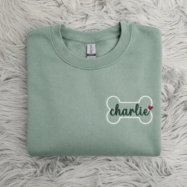 Folded oatmeal sand beige fleece crewneck sweatshirt featuring custom embroidered white dog bone outline with navy cursive pet name on grey fur rug