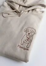 Folded sand beige pullover hoodie featuring custom brown thread embroidered puppy dog portrait and personalized pet name Finnigan underneath