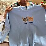 Customer holding up a light blue embroidered crewneck sweatshirt with two cat face portraits named Vita and Tigger