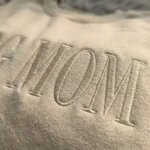 Extreme close-up macro view of dense high-sheen satin stitch embroidery detailing raised letters MOM in ivory thread on sand fleece fabric