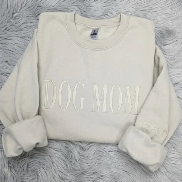 Folded sand beige fleece crewneck sweatshirt featuring elegant tone-on-tone ivory DOG MOM embroidery and personalized pet name Charlie with paw print on sleeve cuff on grey fur