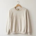 Sand beige crewneck sweatshirt on wooden hanger against white plaster wall showcasing full front DOG MOM embroidery and wrist cuff customization