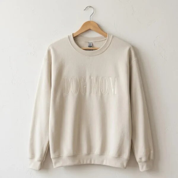 Sand beige crewneck sweatshirt on wooden hanger against white plaster wall showcasing full front DOG MOM embroidery and wrist cuff customization