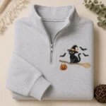 Folded ash grey quarter zip fleece pullover featuring embroidered black cat in witch hat riding a broomstick with jack-o-lantern pumpkin candy bucket and flying bats