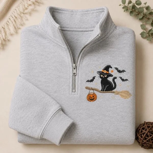 Folded ash grey quarter zip fleece pullover featuring embroidered black cat in witch hat riding a broomstick with jack-o-lantern pumpkin candy bucket and flying bats
