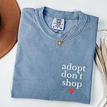 Blue Jean T-shirt laid flat with a necklace and a felt hat, embroidered with adopt don't shop in white stacked letters and a red heart