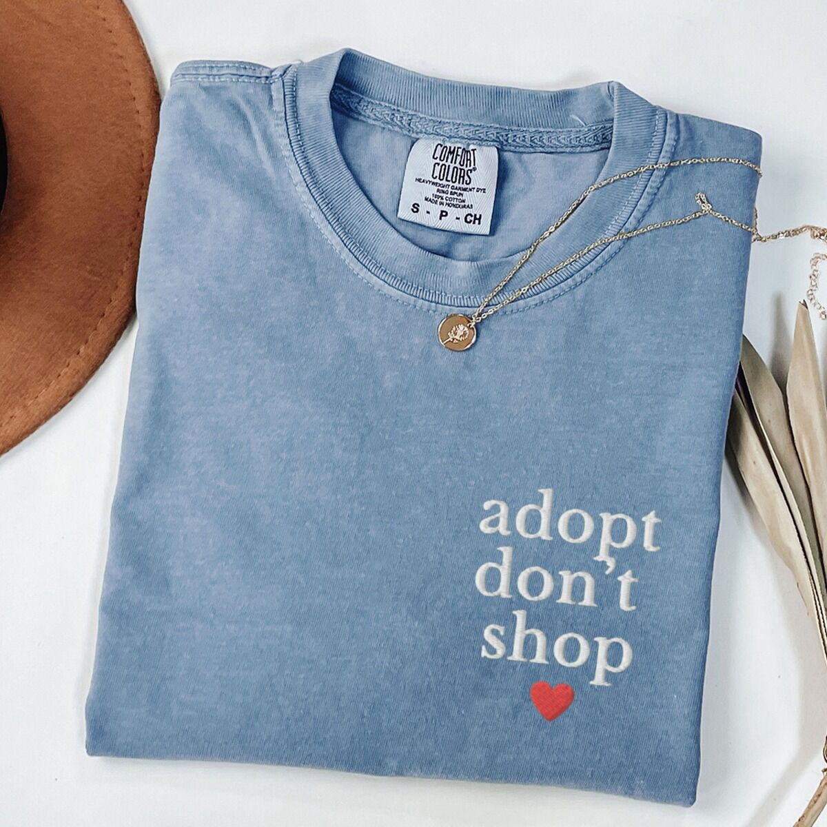 Adopt Don't Shop Shirt – Blue Jean Blue Jean T-shirt laid flat with a necklace and a felt hat, embroidered with adopt don't shop in white stacked letters and a red heart