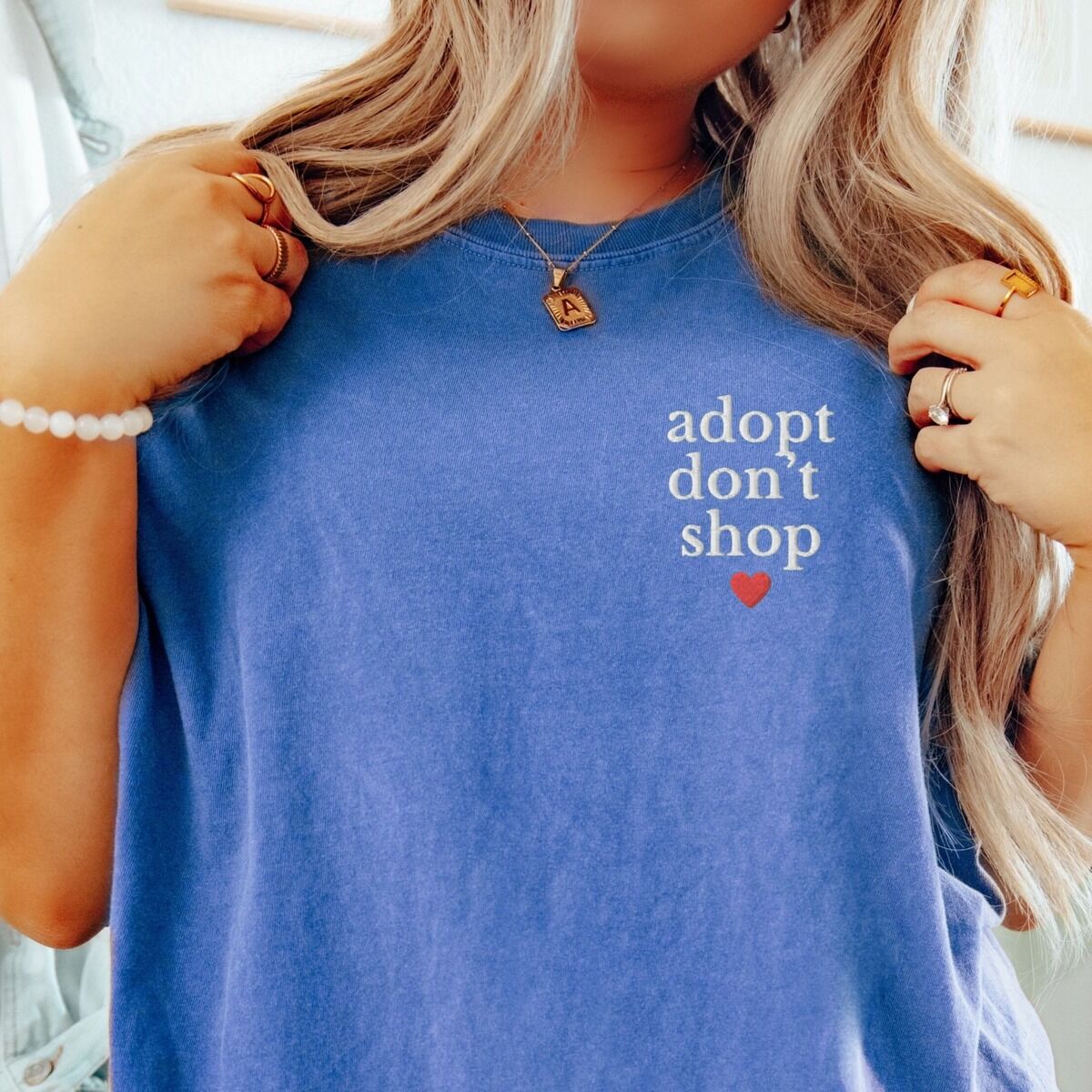 Adopt Don't Shop Shirt – Flo Blue Woman in a Flo Blue T-shirt embroidered with adopt don't shop in white stacked letters and a small red heart on the left chest