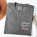 Grey T-shirt laid flat with a necklace and a felt hat, embroidered with adopt don't shop in white stacked letters and a small red heart