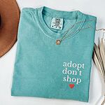 Seafoam T-shirt laid flat with a necklace and dried palm leaves, embroidered with adopt don't shop in white stacked letters and a red heart