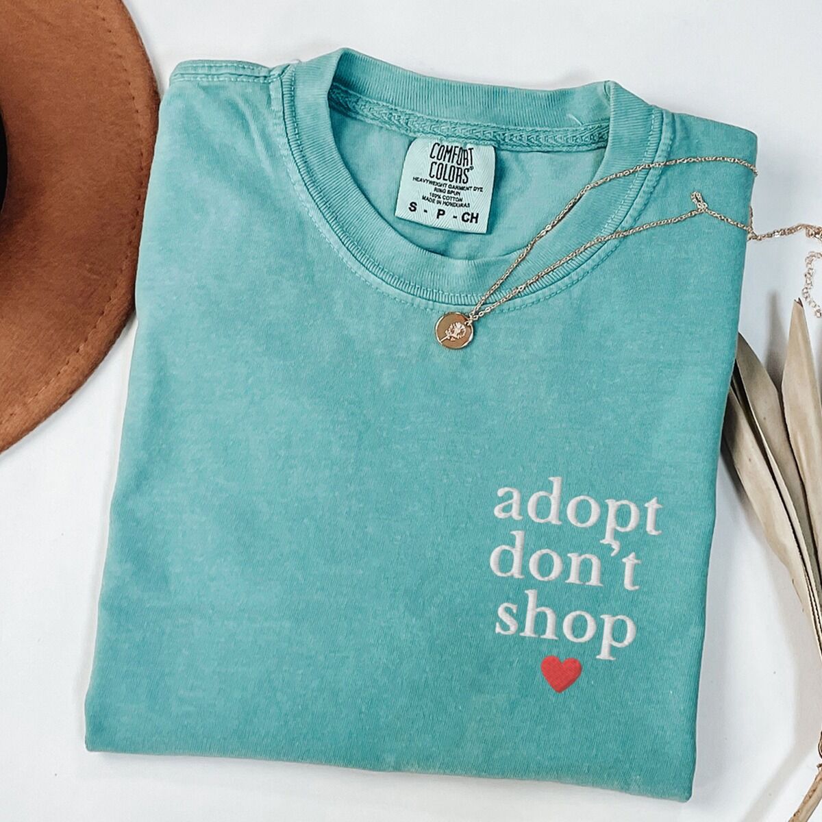 Adopt Don't Shop Shirt – Seafoam Seafoam T-shirt laid flat with a necklace and dried palm leaves, embroidered with adopt don't shop in white stacked letters and a red heart