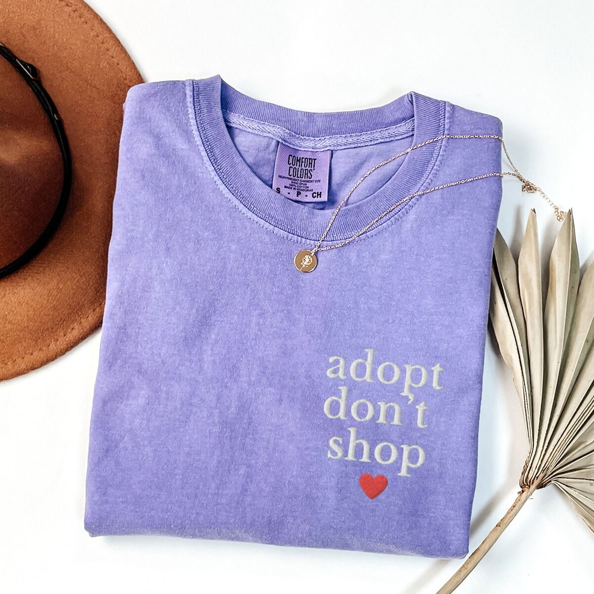 Adopt Don't Shop Shirt – Violet Violet T-shirt laid flat with a necklace and dried palm leaves, embroidered with adopt don't shop in white stacked letters and a red heart