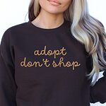 Woman in a dark crewneck sweatshirt embroidered with adopt don't shop in a gold cursive script across the chest