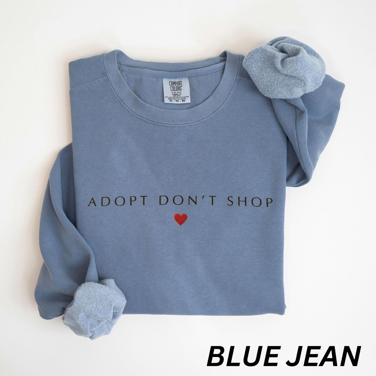 Adopt Don't Shop Sweatshirt – Blue Jean Blue Jean crewneck sweatshirt laid flat on white, embroidered with ADOPT DON'T SHOP in dark spaced capitals and a red heart