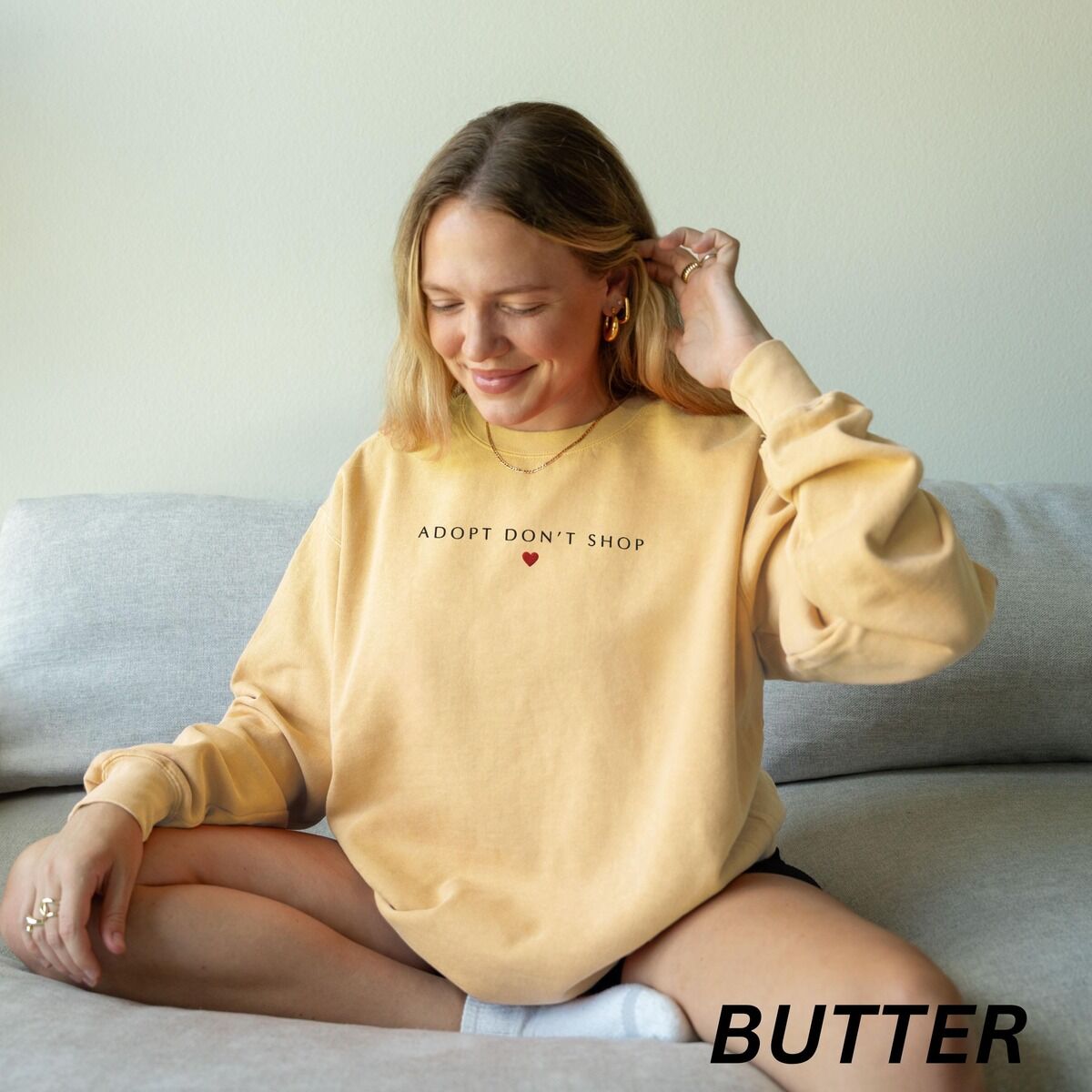 Adopt Don't Shop Sweatshirt – Butter Woman sitting on a sofa in a Butter crewneck sweatshirt embroidered with ADOPT DON'T SHOP in dark capitals and a small red heart